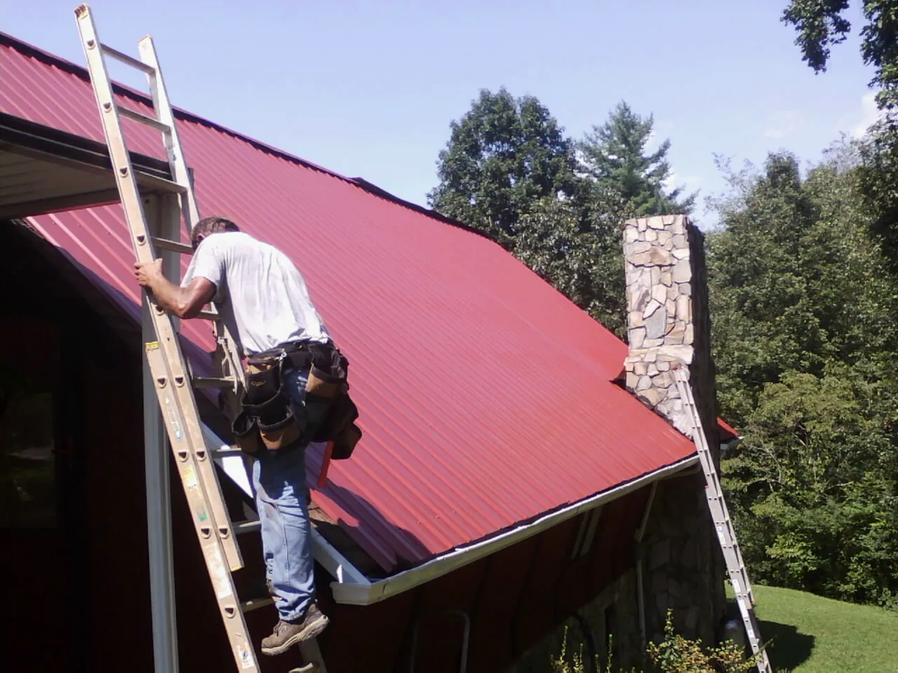Roofer on a ladder beside a red metal roof and stone chimney — Soffit & Fascia Repair by Roofing Brothers in Duncansville